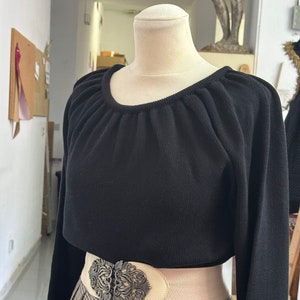 May include: A black long-sleeved sweater with a gathered neckline and a white elastic belt with a large ornate silver buckle. The sweater is worn over a plaid skirt.