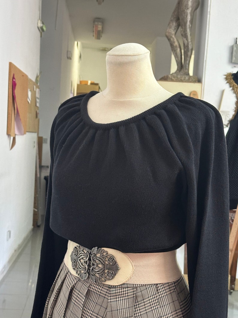 May include: A black long-sleeved sweater with a gathered neckline and a white elastic belt with a large ornate silver buckle. The sweater is worn over a plaid skirt.