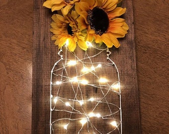 May include: A wooden sign with a mason jar shape outlined with fairy lights and a bouquet of yellow sunflowers.