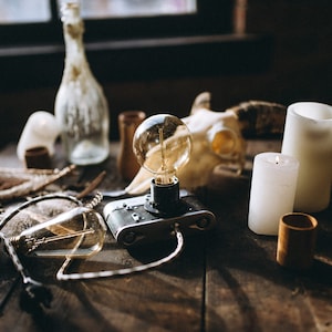 Puede incluir: Una cámara vintage con una bombilla conectada, sentada en una mesa de madera con un cráneo, velas y otros objetos decorativos.