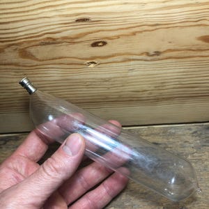 Könnte beinhalten: Eine klare Glasampulle mit einem silberfarbenen Metallverschluss. Die Ampulle wird in einer Hand vor einem Holzgrund gehalten. Die Ampulle ist zylindrisch mit einem abgerundeten Ende und einer Spitze.