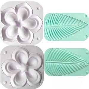 Puede incluir: Dos moldes de silicona blancos para hornear. Un molde tiene forma de flor con cinco p&eacute;talos. El otro molde tiene forma de hoja con un patr&oacute;n de nervaduras detallado.