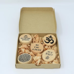 Könnte beinhalten: Ein Set aus fünf Holz-Untersetzern in einer Kartonbox. Die Untersetzer zeigen inspirierende Botschaften und Symbole, darunter "We Rise By Lifting Others", "Be The Light", "Find The Beauty In Every Day", "Om" und ein Lebensblume-Design.