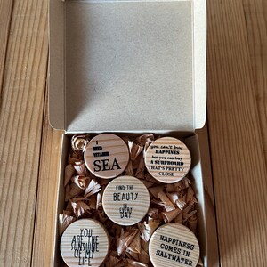 Könnte beinhalten: Eine Holzkiste gefüllt mit Holzspänen enthält sechs runde Holzscheiben mit inspirierenden Zitaten. Die Zitate lauten: "Ich brauche Vitamin MEER", "Glück kann man nicht kaufen, aber man kann ein Surfbrett kaufen, das ist schon ziemlich nah dran", "Finde die Schönheit in jedem Tag", "Du bist der Sonnenschein meines Lebens", und "Glück kommt im Salzwasser".