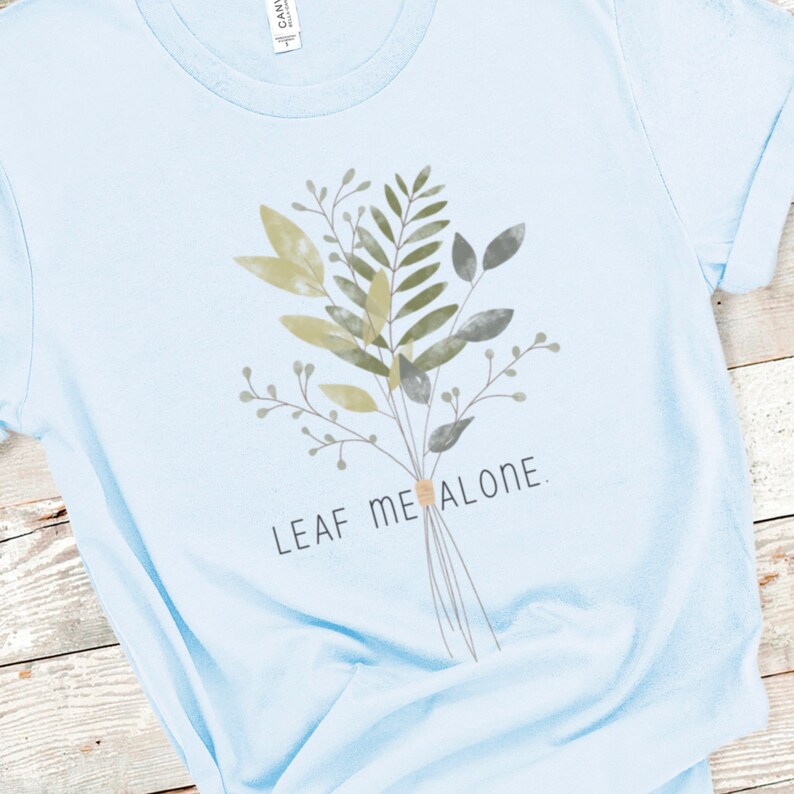 May include: Light blue t-shirt with a watercolor-style illustration of a bouquet of green and brown leaves. The text "LEAF ME ALONE" is printed below the bouquet.