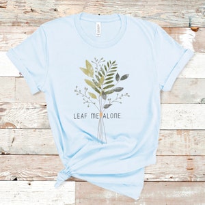 May include: Light blue t-shirt with a watercolor-style illustration of a bouquet of green leaves and the text "LEAF ME ALONE."