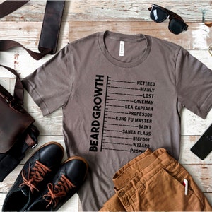 May include: A gray t-shirt with a black graphic of a beard growth chart. The chart lists different stages of beard growth, including "Retired", "Manly", "Lost", "Caveman", "Sea Captain", "Professor", "Kung Fu Master", "Saint", "Santa Claus", "Bigfoot", "Wizard", and "Prophet".