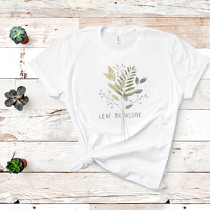 May include: White t-shirt with a watercolor illustration of a bouquet of green and gray leaves. The text "LEAF ME ALONE." is printed below the bouquet.