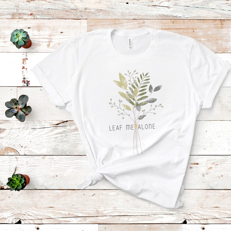 May include: White t-shirt with a watercolor illustration of a bouquet of green and gray leaves. The text "LEAF ME ALONE." is printed below the bouquet.