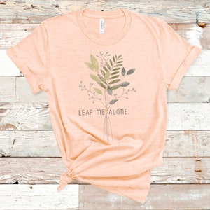 May include: A peach-colored t-shirt with a watercolor-style floral design and the text "LEAF ME ALONE."
