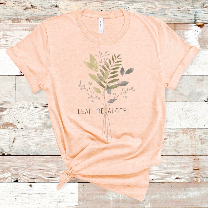 May include: A peach-colored t-shirt with a watercolor-style floral design and the text "LEAF ME ALONE."