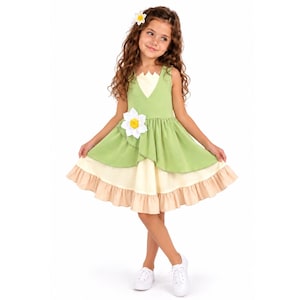 Peut inclure: Une robe sans manches avec un corsage vert clair et une jupe superposée composée d'une section centrale jaune pâle et d'un ourlet beige à volants. La robe est ornée d'applications de fleurs blanches sur le corsage et dans les cheveux, avec des baskets blanches.