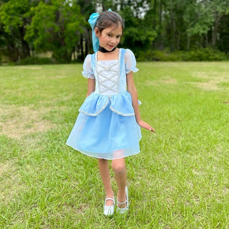 May include: A light blue princess dress with a square neckline, puff sleeves, and a layered skirt. The bodice features white crisscross detailing and silver trim. The outfit is completed with a matching blue bow and silver shoes.