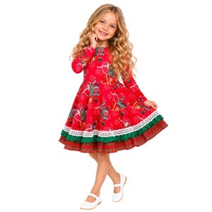 May include: A long-sleeved red dress featuring a repeating nutcracker and snowflake print. The skirt has a white lace trim, a solid green ruffled layer, and a red and green plaid hem. The dress has a round neckline and a flared silhouette.