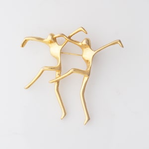 May include: A gold-toned brooch featuring two stylized figures in a dancing pose. The figures have outstretched arms and bent legs, creating a dynamic and artistic design. The brooch is secured with a pin on the back.