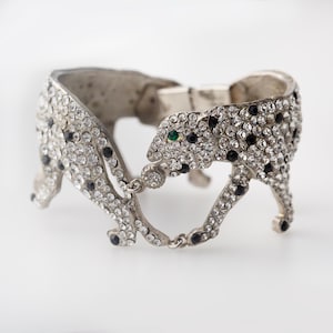 May include: A silver-toned hinged cuff bracelet featuring two stylized leopard figures facing each other. The leopards are encrusted with clear and black rhinestones, with a single green rhinestone eye visible on one of the leopard heads.