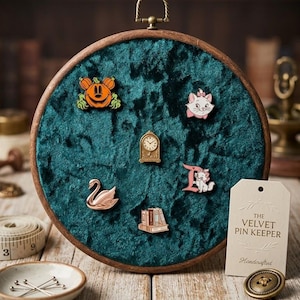 May include: A circular wooden embroidery hoop frame containing dark teal crushed velvet fabric, used as a pin display board. It features five enamel pins: a pumpkin, a cat, a clock, a swan, and books. A tag reads THE VELVET PIN KEEPER.