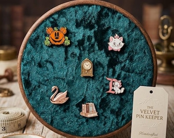 May include: A circular wooden embroidery hoop frame containing dark teal crushed velvet fabric, used as a pin display board. It features five enamel pins: a pumpkin, a cat, a clock, a swan, and books. A tag reads THE VELVET PIN KEEPER.