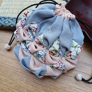 Könnte beinhalten: Ein runder Stoffbeutel mit Kordelzug in Blau-, Rosa- und Blumenmustertönen. Der Beutel besteht aus weichem Stoff und hat einen schwarzen Kordelzugverschluss mit kleinen, weißen Pompons. Er liegt auf einer Holzoberfläche.