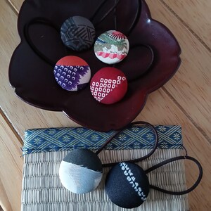 May include: A collection of fabric-covered hair ties with various patterns, including geometric and floral designs. The hair ties are in a dark brown decorative dish and on a woven mat with blue trim. The colors include red, white, blue, and black.