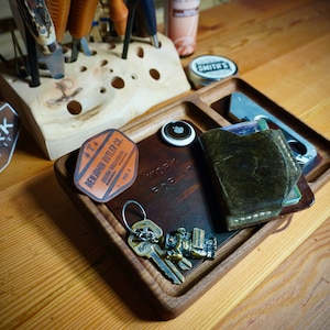 May include: A wooden tray with a leather inlay, featuring the text "Work for it". The tray contains a small leather wallet, a keychain with keys and a small metal figure, a round metal disc, and a leather patch with the text "Belmont Butler Co.".