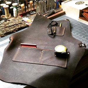 May include: A brown leather apron with multiple pockets, including one with a yellow tape measure and another with a red tool. The apron is lying on a black and gray cutting mat.