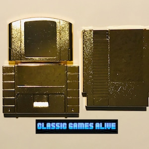 以下が含まれることがあります： 金色のビデオゲームカートリッジが2つ並んでいます。片方のカートリッジは上端が丸みを帯びており、もう片方は長方形で側面に溝があります。カートリッジの下には「CLASSIC GAMES ALIVE」という青い文字が印刷されています。