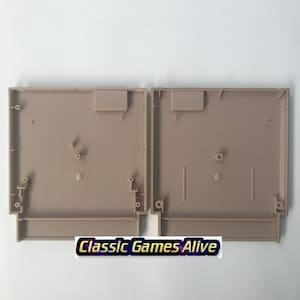 May include: Two tan-colored, empty Nintendo Entertainment System (NES) cartridge shells. Each shell has a rectangular shape with various internal features, including posts and slots. The text "Classic Games Alive" is visible at the bottom.