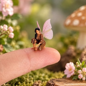 May include: A miniature figurine of a fairy with dark hair, wearing a brown outfit, and featuring translucent pink wings. The fairy is in a kneeling position, interacting with a small brown pot, and is balanced on the tip of a human finger.