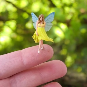 May include: A miniature fairy figurine with a yellow dress and blue wings. The fairy is standing on a finger, with a blurred green background. The fairy has brown hair and is wearing small, white shoes.