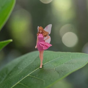 Op de afbeelding: Een miniatuur feeënbeeldje met een roze jurk en doorschijnende vleugels staat op een groot groen blad. De fee heeft rood haar en poseert met één hand op haar heup. De achtergrond is wazig met zachte groene en witte bokeh.