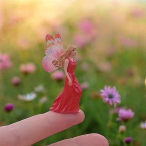 May include: A miniature fairy figurine with a red dress and butterfly wings. The fairy has long blonde hair and is standing on a fingertip. The background is a blurred field of pink and white flowers.