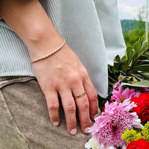May include: A gold bracelet and ring are worn on a person's hand, with a bouquet of colorful flowers in the background. The bracelet is a thin, beaded design. The ring is a simple band. The flowers include pink, red, yellow, and green.