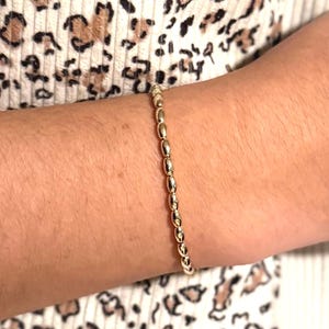 以下が含まれることがあります： 楕円形のビーズが付いたゴールドのブレスレットが手首に着用されています。ブレスレットは、小さな光沢のあるビーズを繋ぎ合わせて作られています。ヒョウ柄のトップスを着ています。