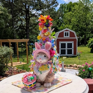 May include: A ceramic Easter bunny figurine with a pink hat and a basket of colorful eggs. The bunny is decorated with flowers and ribbons. The figurine is sitting on a white table with a checkered tablecloth. The background is a blurry image of a garden with trees and a red shed.