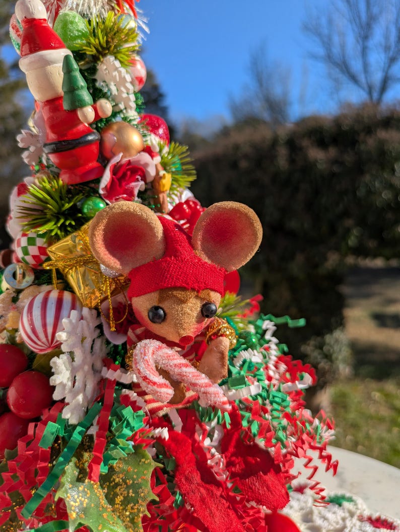 May include: A miniature Christmas tree decorated with red and green tinsel, ornaments, and a small mouse figurine holding a candy cane.