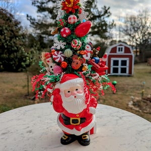 May include: A festive Christmas tree figurine with a Santa Claus base. The tree is adorned with colorful ornaments, red and green tinsel, and a red flower at the top. The Santa figure is wearing a red suit with a black belt and boots.