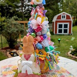 以下が含まれることがあります： パステルカラーの花、卵、リボンで飾られたカラフルなイースターツリーのセンターピース。2羽の陶器製のウサギが木の根元に立っており、編み物のプレースマットの上に置かれています。