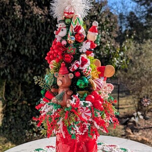 May include: A miniature Christmas tree decorated with red and green ornaments, candy canes, and a small teddy bear. The tree is in a red pot and sitting on a white crocheted doily.