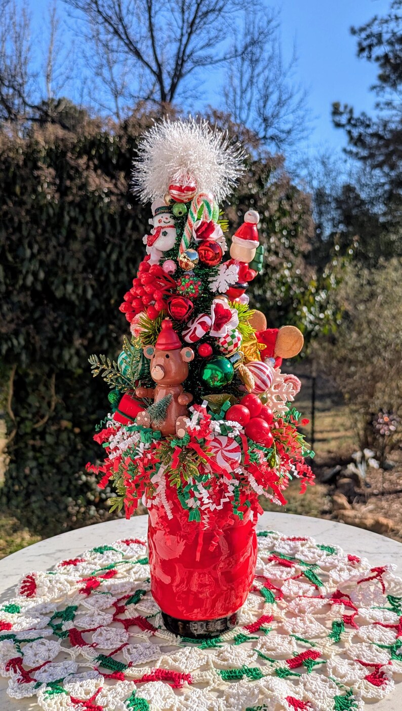 May include: A miniature Christmas tree decorated with red and green ornaments, candy canes, and a small teddy bear. The tree is in a red pot and sitting on a white crocheted doily.