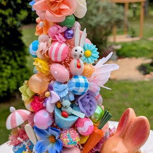 以下が含まれることがあります： パステルピンクのイースターツリーは、カラフルな卵、花、ウサギの置物で飾られています。ツリーは、パステルカラーの卵、花、小さなウサギの置物など、イースターをテーマにしたさまざまな装飾で飾られています。ツリーは、カラフルなイースターグラスで囲まれています。