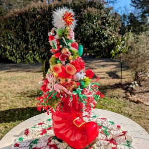 May include: A red ceramic boot with a Christmas tree inside. The tree is decorated with red and green ornaments, candy canes, and a small mouse figurine. The boot is sitting on a white crocheted doily.