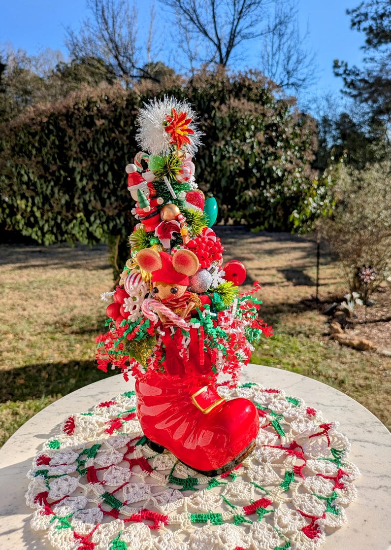 May include: A red ceramic boot with a Christmas tree inside. The tree is decorated with red and green ornaments, candy canes, and a small mouse figurine. The boot is sitting on a white crocheted doily.