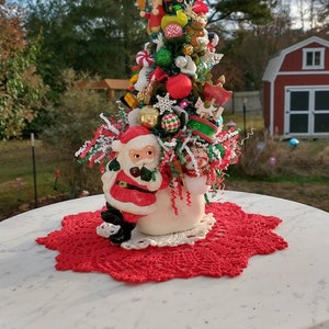 May include: A miniature Christmas tree decorated with ornaments, candy canes, and a Santa Claus figurine. The tree is in a white planter and sits on a red crocheted doily on a white marble table.