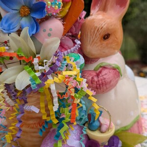 以下が含まれることがあります： カラフルなイースターエッグ、花、リボンでいっぱいのバスケットを持ったセラミック製のウサギの置物。ウサギはピンクのドレスを着ており、茶色の毛皮をしています。バスケットは、かぎ針編みの表面の上に置かれています。