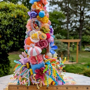 以下が含まれることがあります： パステルカラーの花、卵、リボンで飾られたカラフルなイースターツリー。ツリーの高さは約15cmで、白とピンクの編み物の上に置かれています。ツリーは、小さなウサギ、ミニチュアの傘、小さな青いワゴンなど、さまざまなイースターをテーマにしたアイテムで飾られています。