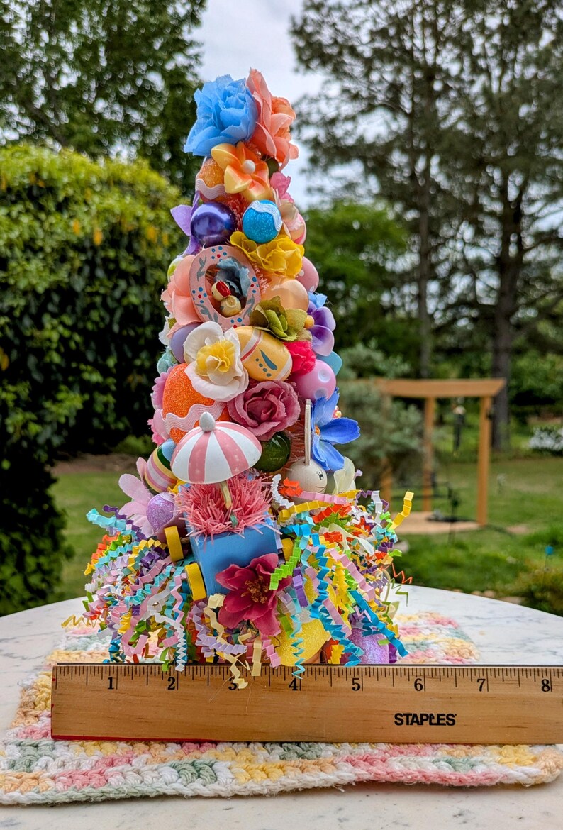 以下が含まれることがあります： パステルカラーの花、卵、リボンで飾られたカラフルなイースターツリー。ツリーの高さは約15cmで、白とピンクの編み物の上に置かれています。ツリーは、小さなウサギ、ミニチュアの傘、小さな青いワゴンなど、さまざまなイースターをテーマにしたアイテムで飾られています。