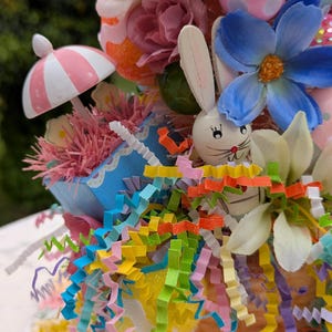 以下が含まれることがあります： カラフルなイースターエッグ、花、木製のウサギが入ったパステルカラーのイースターバスケット。バスケットはピンクと白のストライプの紙で飾られ、カラフルなカーリーペーパーストリームで囲まれています