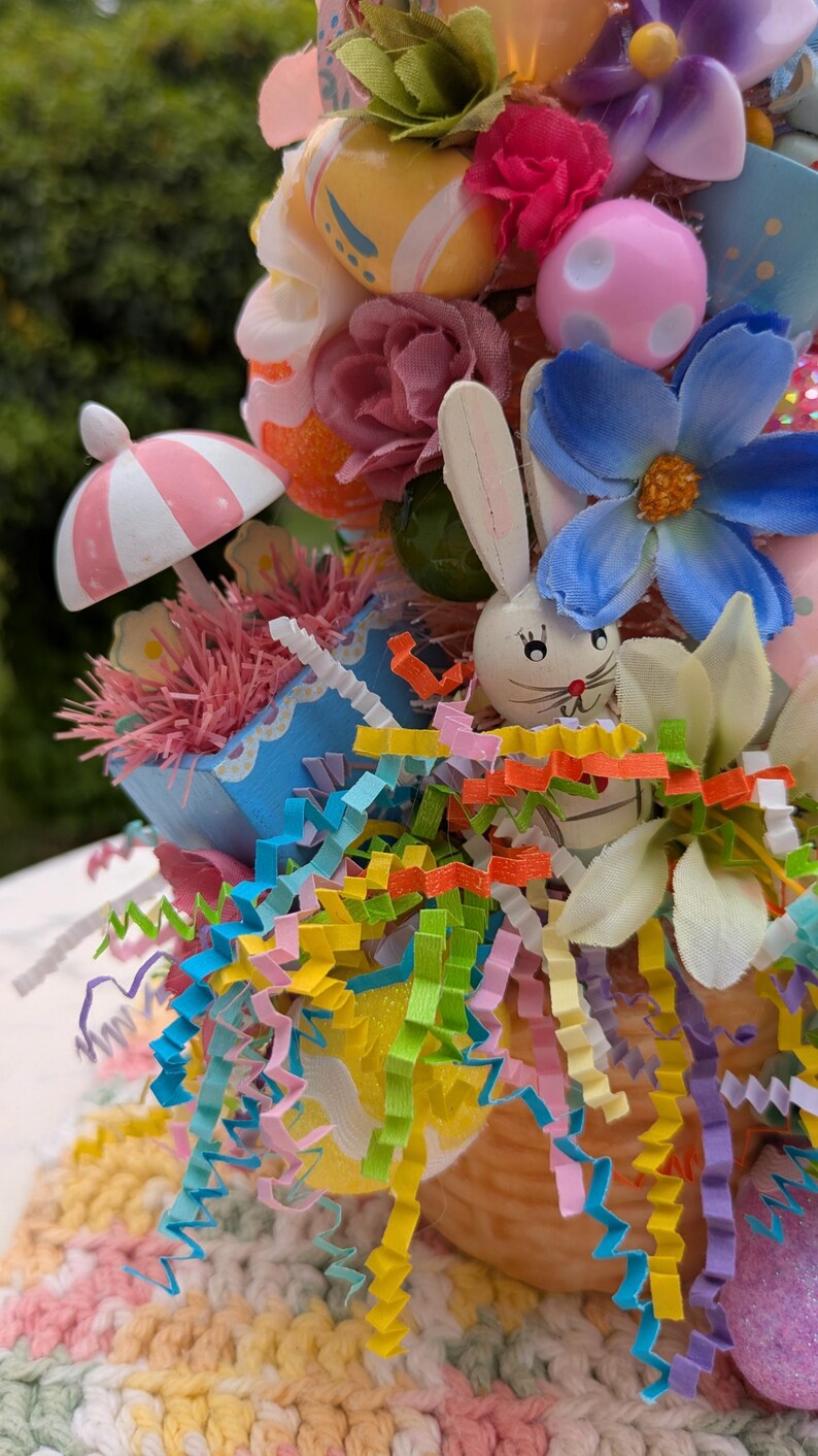 以下が含まれることがあります： カラフルなイースターエッグ、花、木製のウサギが入ったパステルカラーのイースターバスケット。バスケットはピンクと白のストライプの紙で飾られ、カラフルなカーリーペーパーストリームで囲まれています