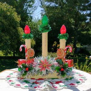 May include: A vintage Christmas candle holder with three lights, decorated with gingerbread men, candy canes, and a poinsettia. The holder is surrounded by silver tinsel and green garland.